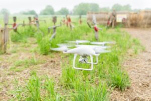 A white drone hovers above a green field, capturing aerial views of the landscape below.